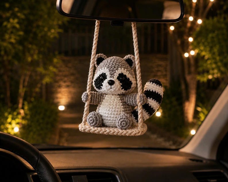 May include: A crocheted raccoon ornament hanging from a rearview mirror. The raccoon is grey, white, and black with a striped tail, sitting on a small, beige crocheted swing suspended by two thick, twisted ropes attached to the mirror mount.