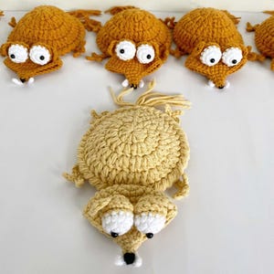 May include: A collection of handmade crochet animal figures in shades of gold and tan. Each figure features a round body, a pointed snout, and two large white circular eyes with black pupils. The figures have small limbs and a tuft of yarn at the back.
