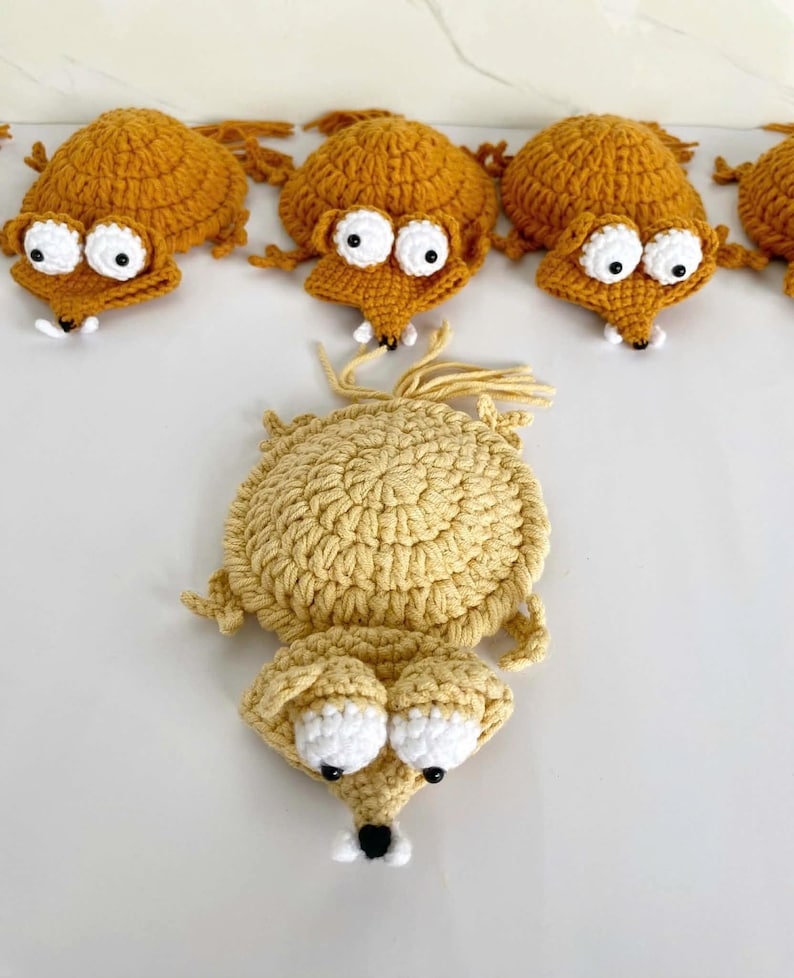 May include: A collection of handmade crochet animal figures in shades of gold and tan. Each figure features a round body, a pointed snout, and two large white circular eyes with black pupils. The figures have small limbs and a tuft of yarn at the back.