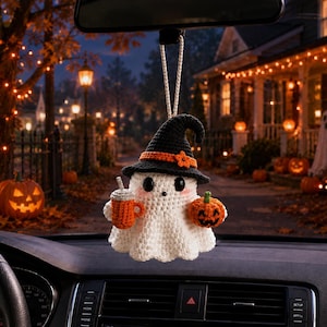 May include: A crocheted ghost ornament hanging from a rearview mirror. The white ghost wears a black witch hat with an orange band and flower, holds an orange mug in one hand, and a small orange jack-o'-lantern in the other hand.