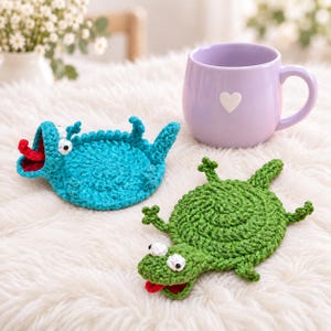 Puede incluir: Dos posavasos de ganchillo hechos a mano con forma de pequeños lagartos, uno en turquesa brillante y otro en verde hierba, cada uno con ojos blancos con pupilas negras y una lengua roja, junto a una taza de cerámica morada con un corazón blanco.