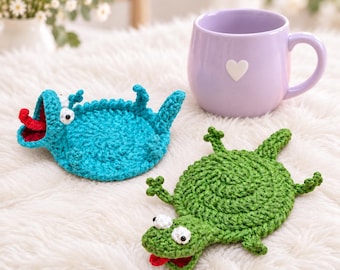 May include: Two handmade crochet coasters shaped like small lizards, one in bright turquoise and one in grass green, each featuring white eyes with black pupils and a red tongue, positioned next to a light purple ceramic mug with a white heart.