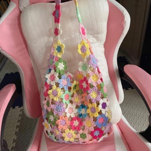 Op de afbeelding: Een handgemaakte gehaakte tas met een bloemenpatroon in verschillende pastelkleuren. De tas is gemaakt van garen en heeft een schouderriem. De bloemen zijn in tinten roze, geel, blauw, groen en wit. De tas ligt op een witte en roze stoel.