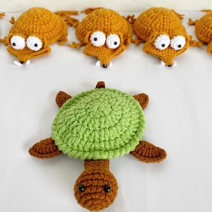 May include: A collection of crocheted items including a green and brown turtle with a textured shell and a brown head, alongside several orange fox-like figures with white eyes, black pupils, and small white teeth arranged in a row above the turtle.
