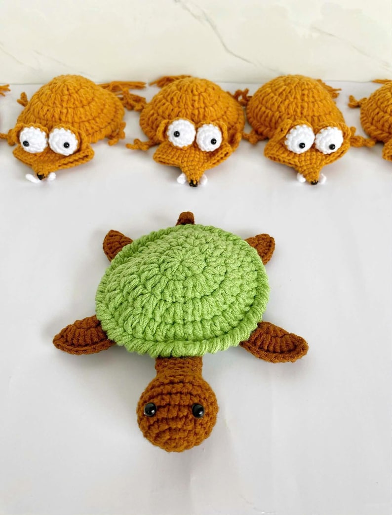 May include: A collection of crocheted items including a green and brown turtle with a textured shell and a brown head, alongside several orange fox-like figures with white eyes, black pupils, and small white teeth arranged in a row above the turtle.
