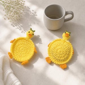 May include: Two round, yellow crocheted coasters shaped like ducks, each featuring a small head with a green tuft, orange beak, and orange feet. The coasters are positioned near a plain, light gray ceramic mug on a textured white fabric surface.