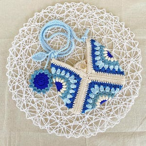 Op de afbeelding: Een gehaakte blauwe en witte handtas met een granny square patroon. De tas heeft een lichtblauwe band en een kleine blauwe bloemenhanger.