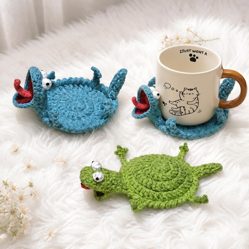May include: Two blue and one green crocheted coasters shaped like creatures with wide mouths, red tongues, and white eyes. One blue coaster supports a cream-colored ceramic mug featuring a black line drawing of a cat and the text I JUST WONT A.