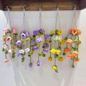 Może przedstawiać: Kolekcja pięciu wiszących koszyków z kwiatami wykonanych na szydełku. Każdy koszyk ma inny kolor, w tym różowy, fioletowy, żółty i pomarańczowy. Kwiaty i pnącza opadają kaskadowo w dół, tworząc dekoracyjną ekspozycję. Koszyki wiszą na drewnianej półce.