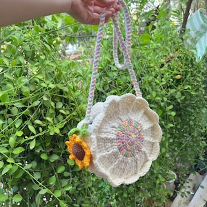 May include: A cream-colored crochet bag with a sunflower design. The bag features a large, round body with a scalloped edge and a colorful floral center. A small, crocheted sunflower is attached to the side, and the strap is a pastel rainbow color.