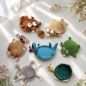May include: A collection of seven crocheted animal-shaped coasters including a brown squirrel, a tan squirrel, a small brown acorn, a light brown koala, a blue creature with a red tongue, a green frog, a gray mouse, and a dark green turtle.