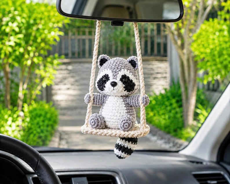 May include: A crocheted raccoon ornament hanging from a rearview mirror. The gray, white, and black raccoon sits on a small, beige crocheted swing suspended by two thick, twisted ropes. The raccoon has a striped tail hanging below the swing.
