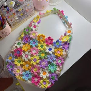 May include: A handmade crochet tote bag featuring a colorful floral design. The bag is crafted with a white base and adorned with vibrant, multi-colored crochet flowers in shades of pink, yellow, blue, and purple. The strap is also decorated with crochet flowers.