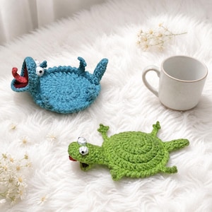 May include: Two crocheted coasters shaped like creatures, one blue with an open mouth and red tongue, and one green with a flat body and protruding eyes. A plain white ceramic mug sits nearby, completing this set of handmade textile drink coasters.