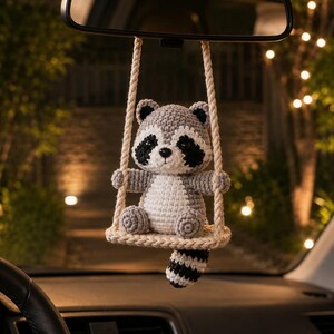 May include: A crocheted raccoon ornament hanging from a rearview mirror. The raccoon is gray, white, and black, sitting on a small, cream-colored crocheted swing suspended by two thick, twisted ropes. The raccoon has a striped tail hanging down.