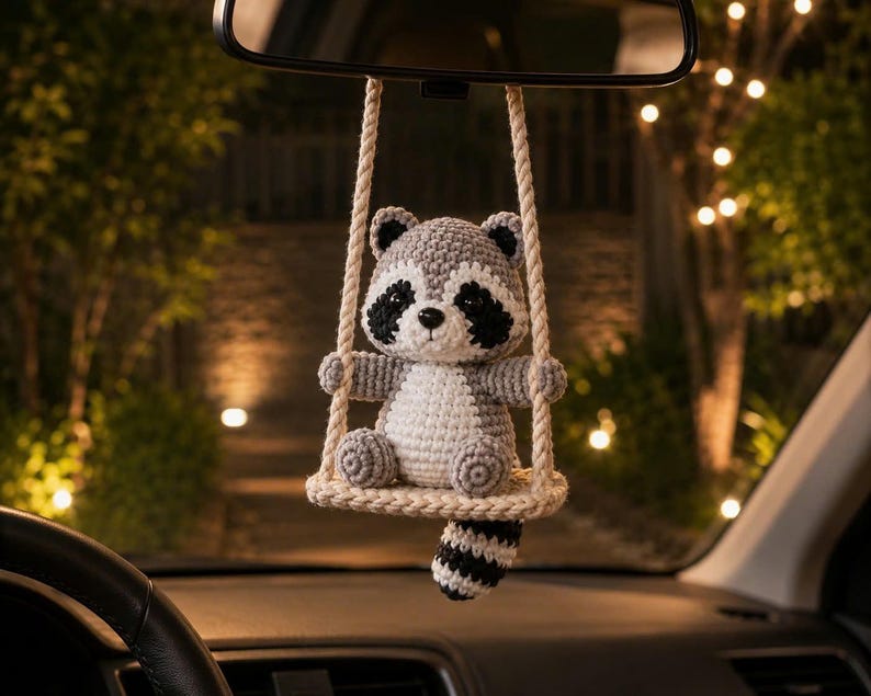 May include: A crocheted raccoon ornament hanging from a rearview mirror. The raccoon is gray, white, and black, sitting on a small, cream-colored crocheted swing suspended by two thick, twisted ropes. The raccoon has a striped tail hanging down.