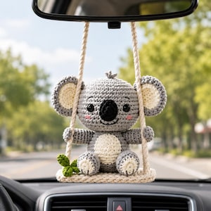 May include: A crocheted koala ornament hanging from a rearview mirror. The gray and cream koala sits on a small cream-colored swing suspended by two braided ropes. A small green four-leaf clover is attached to the side of the swing seat.