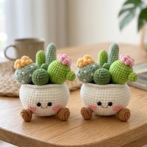 May include: Two handmade crochet cactus plants in white pots with smiling faces and small brown feet. The cacti are green with pink and yellow flower accents, set on a wooden table. The background includes a neutral-colored mug and a plant.