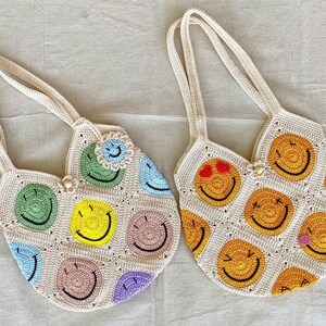 May include: Two crocheted tote bags with smiley face designs. One bag is cream with multi-colored smiley faces and the other is cream with yellow smiley faces.