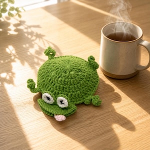 May include: A round, green crocheted frog coaster featuring a textured surface, two white eyes with black pupils, and a small pink tongue protruding from the mouth. The frog has four small limbs extending from the sides of its circular body.