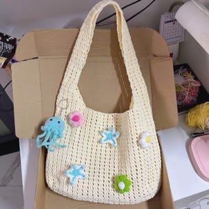May include: A cream-colored crochet tote bag with a long strap. The bag is decorated with crochet starfish, flowers, and a blue jellyfish charm. The bag is sitting inside a cardboard box.