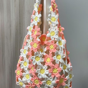 Op de afbeelding: Een handgemaakte gehaakte tas met een bloemenpatroon. De tas is gemaakt van wit, geel en roze garen, met een trekkoordsluiting en schouderbanden. De tas is ongeveer 30 cm breed en 35 cm hoog.