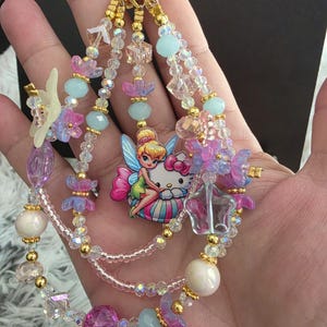 May include: A beaded keychain featuring a central charm of a fairy character sitting on a cat character. The design includes multiple strands of clear, pink, purple, and light blue beads, interspersed with gold-toned spacers and a gold metal ring.