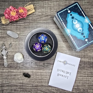 May include: A set of three colorful dice with gold numbers in a black bowl. The dice are blue, purple, and green. The bowl is on a wooden surface with a deck of tarot cards, a booklet titled "Astro Dice Booklet", and other items.