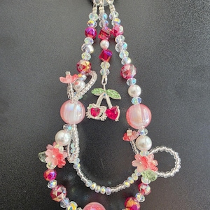 May include: A beaded keychain featuring a silver-toned ring, clear iridescent beads, pink spherical beads, and small red faceted crystals. The design includes floral-shaped accents and a central silver-toned charm shaped like two cherries.