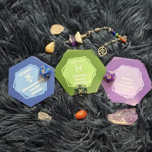 May include: An assortment of crystals, dice, and cards with astrological symbols and text. The cards are blue, green, and purple. The dice are blue, green, and purple. A beaded bracelet with colorful beads and a charm is also present.