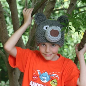 May include: A child wearing a gray crocheted bear hat with brown felt nose and ears. The child is wearing an orange t-shirt with a cartoon graphic of a fish and the words "Angry Birds".