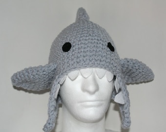 May include: Gray crocheted shark hat with black eyes and white felt teeth. The hat has long ear flaps and is worn on a white mannequin head.