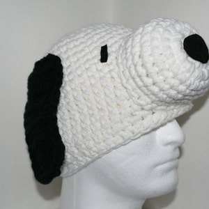 May include: White crocheted hat with black felt ears and nose, resembling a cartoon dog. The hat is worn on a white mannequin head.