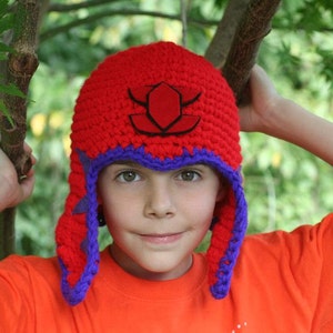May include: A child wearing a red and purple crocheted hat with a black and red emblem. The hat has a long flap that covers the ears and neck.