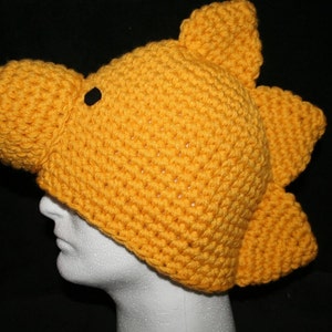 May include: A handmade, yellow crochet hat shaped like a dinosaur's head. The hat features a rounded snout, a black eye, and three dorsal fins. The hat is designed to fit a human head.