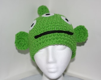 May include: Green crocheted hat with three eyes and a black mouth, resembling an alien character. The hat has two green crocheted ears.