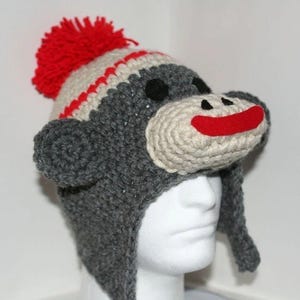 May include: A handmade crochet hat in the shape of a monkey. The hat is primarily gray with a white face and red accents. It features black eyes, a red mouth, and a red pom-pom on top. The hat has ear flaps.