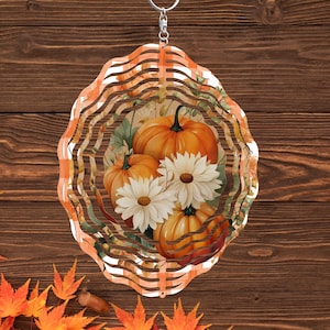 May include: A decorative, layered metal wind spinner featuring a fall theme. The design includes orange pumpkins and white daisies, set against a backdrop of green leaves. The outer frame has an orange and white striped pattern.
