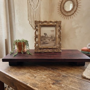 May include: A rectangular dark brown wooden riser with four short feet, featuring a gold ornate picture frame containing a desert landscape print and a small terracotta pot with a trailing green plant placed on its surface.