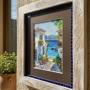 May include: A square wall art piece featuring a textured painting of a Mediterranean coastal scene with white buildings and blue sea. The artwork is encased in a light-colored, distressed wooden frame with carved floral details and a blue mosaic border.