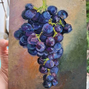 May include: An oil painting on a rectangular canvas depicting a cluster of dark purple and blue grapes with visible green stems. The grapes are rendered with thick, textured brushstrokes against a muted, earthy background of brown and green tones.