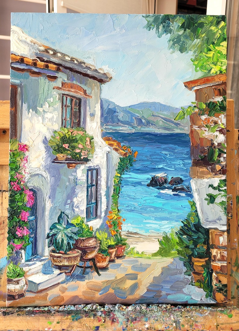 A painting of a white building with a blue door and windows, overlooking a blue ocean with a rocky shore. The building is surrounded by green plants and flowers.