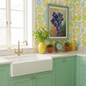 May include: A kitchen featuring mint green cabinets, a white farmhouse sink with a brass faucet, and a yellow vase with a green plant. The wall has fruit-patterned wallpaper and a framed oil painting of a bunch of dark purple grapes.