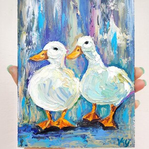 Oil painting of two white ducks with orange beaks and feet. The ducks are painted against a background of blue, purple, and teal brushstrokes. The painting is held by a person.