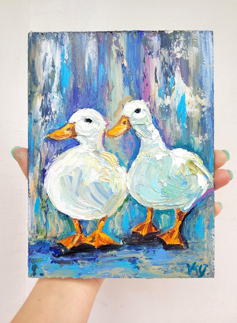 Oil painting of two white ducks with orange beaks and feet. The ducks are painted against a background of blue, purple, and teal brushstrokes. The painting is held by a person.