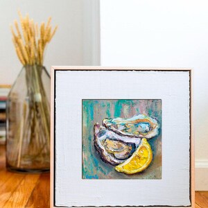 May include: Oil painting of oysters and a lemon slice. The artwork features a square canvas with a white border and a light brown frame. The painting has a teal and brown background with the oysters and lemon in the foreground.