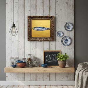 May include: A framed painting of a fish on a gold background hangs on a white wood-paneled wall. Decorative blue and white plates are mounted nearby. A shelf below holds jars, bowls, and a chalkboard that reads "CATCH".
