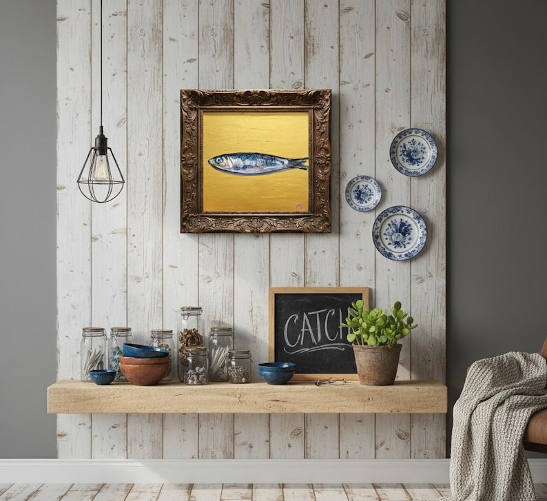 May include: A framed painting of a fish on a gold background hangs on a white wood-paneled wall. Decorative blue and white plates are mounted nearby. A shelf below holds jars, bowls, and a chalkboard that reads "CATCH".