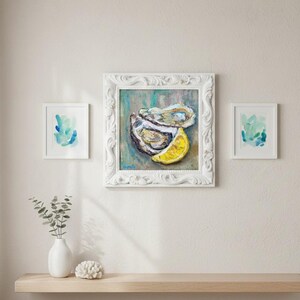 May include: A white framed painting of an oyster and a lemon slice. Two smaller framed abstract watercolor paintings are on either side. A white vase with greenery and a white coral-like object sit on a wooden shelf below.