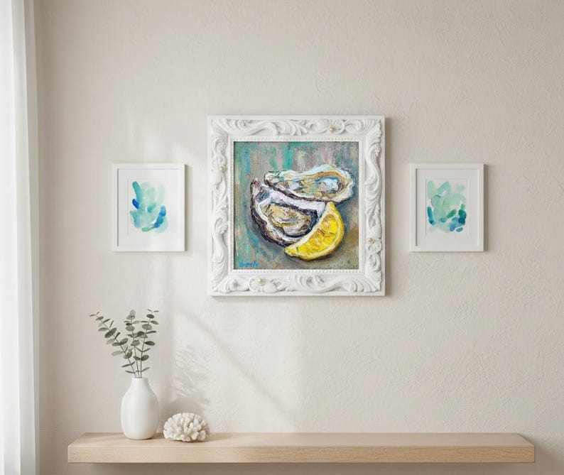 May include: A white framed painting of an oyster and a lemon slice. Two smaller framed abstract watercolor paintings are on either side. A white vase with greenery and a white coral-like object sit on a wooden shelf below.
